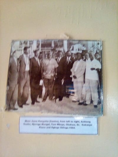 This image depicts a faded, slightly discolored black and white photograph mounted on a light beige wall. The photograph is enclosed in a clear protective sheet, adhered to the wall with a small piece of white tape visible above its center. The physical photograph is located in the city of South Sakwa ward, Kenya.
The photograph itself shows a group of approximately eight to nine men standing together outdoors in what appears to be an urban setting, with indistinct architectural structures in the background. The men are arranged in a horizontal line, facing generally towards the viewer. Most of the men are wearing suits, jackets, and trousers. In the center, Mzee Jomo Kenyatta is identifiable, wearing a lighter, patterned short-sleeved shirt and trousers, and holding a distinctive dark staff in his left hand. To his immediate right (viewer's left), a man in a dark suit stands. Further to the viewer's left are three other men in suits. To Kenyatta's immediate left (viewer's right), a man in a light-colored suit or traditional attire is visible, and further to his left, at least two more men in dark suits are present. The individuals are identified by a caption positioned directly below the photograph.
The caption, printed on a white rectangular label, reads: "Mzee Jomo Kenyatta (Centre), from left to right, Achieng Oneko, Njoroge Mungai, Tom Mboya, Chokwe, Dr. Gakonyo Kiano and Oginga Odinga. 1964." This identifies the central figure as Mzee Jomo Kenyatta and lists other prominent Kenyan figures from the specified year. The photograph exhibits signs of age, including blurring, fading, and a slight blue tint along its right edge.