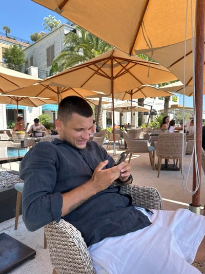 A man sits in a woven chair outdoors, holding a smartphone and looking at it with a strained expression. He is wearing a dark blue, collared shirt and white shorts. The background shows a patio area with multiple large, tan umbrellas providing shade over tables and chairs. Several other people are visible in the distance, seated at tables. Behind the patio, there is a building with white walls and dark shutters, surrounded by palm trees and other greenery. The sky is clear and blue. The scene appears to be a public outdoor seating area, likely a restaurant or cafe, in Donja Lastva, Montenegro.