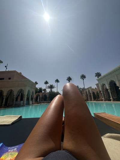 The image shows the legs of a person lying on a lounge chair by a swimming pool in the city of Ouled Jelal, Morocco. The sun is high in the sky and the sky is clear blue. There are palm trees on either side of the pool. The person is wearing a black bathing suit and a book titled "Prakriti" is lying on the lounge chair. The pool is surrounded by a low wall with arched openings. The building in the background has white walls and a brown roof. The person is facing the sky and looking up. The pool water is a vibrant turquoise color. The lounge chair is made of dark fabric and wood. The environment is tropical and the weather appears to be pleasant. The view from the lounge chair is of the pool, palm trees, and a building in the background. The picture was likely taken in the afternoon during the summer.