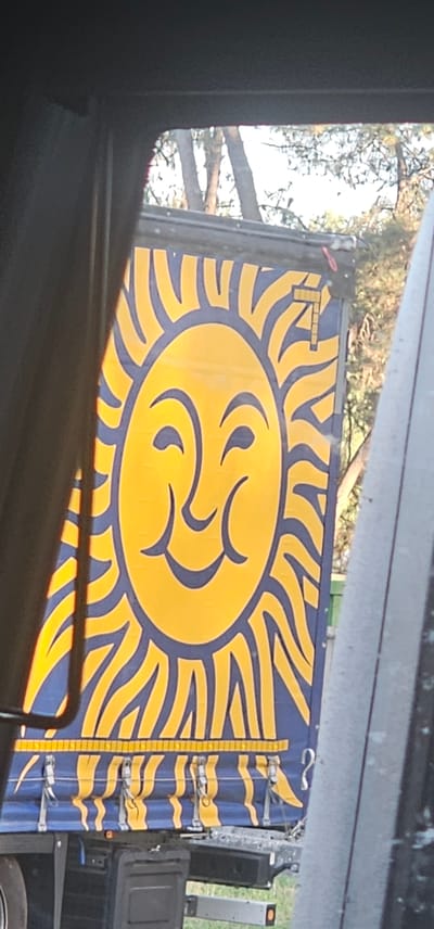 A close-up, eye-level shot captures the side of a blue truck trailer adorned with a large, stylized yellow sun with a smiling face. The sun's rays are depicted as wavy yellow lines outlined in blue, radiating outward from its center. The trailer is partially obscured by the interior of a vehicle, suggesting the photo was taken from a car. The background shows trees and foliage, indicating an outdoor setting. The truck appears to be stationary, possibly parked along a roadside in Cestas, France.