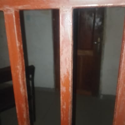 The image shows a view through a set of orange bars, likely part of a gate or barrier. In the background, a dark wooden door with a silver handle is visible, suggesting an interior space. To the left, a portion of a dark chair or bench can be seen. The lighting is dim, and the overall atmosphere appears somewhat aged or neglected, with some wear visible on the orange bars. The scene is not clearly identifiable as being in Ogbomosho, Nigeria, as there are no specific landmarks or cultural indicators present.