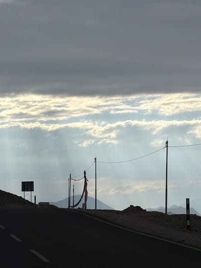 This image captures a desolate mountain road in Chumkar, India, under a dramatic, cloudy sky. The foreground is dominated by a dark asphalt road with white dashed lines. To the left, a hill rises, topped with a silhouette of a signpost. Further along the road, several utility poles stand, one strung with prayer flags that flutter in the wind. In the distance, a chain of muted mountain ranges stretches across the horizon, partially obscured by haze and the low-hanging clouds. The overall impression is one of solitude and vastness.