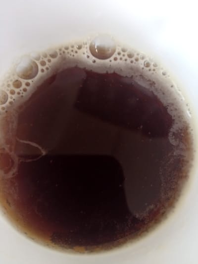 The image is a close-up, overhead view of a dark liquid, likely coffee, in a white cup. The surface of the liquid is covered with a layer of light brown foam and small bubbles. The lighting is bright, causing some glare on the bubbles. There are no people or any identifiable landmarks of Terebovlia, Ukraine, present in the image. The focus is entirely on the beverage and its foamy surface.