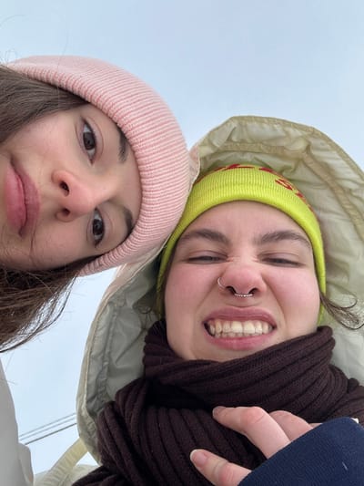 This image depicts a close-up, upward-angled selfie of two individuals, identified as being in Synelnykove, Ukraine. The subject on the left, partially visible, has long dark hair and wears a light pink ribbed beanie, with a neutral expression looking towards the camera. The subject on the right, more fully visible, wears a bright lime green beanie with red patterns, a light-colored hooded jacket, and a dark brown ribbed scarf. This individual exhibits a scrunched facial expression, showing upper teeth, and has a silver beaded septum piercing. A hand, likely belonging to the second individual and wearing a dark blue sleeve, is visible near her neck, possibly adjusting the scarf. The background is an undifferentiated light grey, suggesting an overcast sky or blurred outdoor environment.