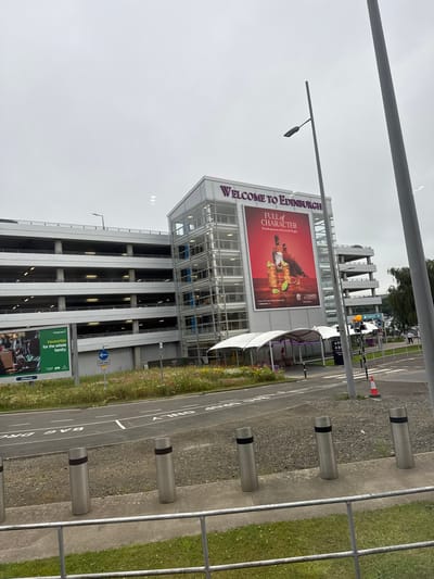 This media file is a photograph taken in the city of Edinburgh, United Kingdom. The image shows a multi-story car park with a large billboard attached to its side. The billboard displays an advertisement for "Edinburgh Airport" with the text "WELCOME TO EDINBURGH" in large purple letters. Below this, it reads "FULL OF CHARACTER" and mentions "Proud sponsors of Scottish Rugby." The advertisement features bottles of amber liquid, limes, and two birds, suggesting a beverage promotion.
The car park itself is modern with a glass facade on the upper levels and open-air levels below. There are several concrete bollards with black bands lining the paved area in front of the car park. To the right of the bollards, there is a grassy area and a metal fence. A road with white lane markings is visible, along with a "One way" sign. Further in the background, more trees and buildings can be seen, along with what appears to be an airport terminal. The sky is overcast, suggesting a cloudy day. The time of day is difficult to determine precisely due to the lighting conditions. No people are prominently featured in the foreground, though some figures are visible in the distance. The overall scene suggests an arrival or departure point, likely an airport due to the "Edinburgh Airport" branding on the billboard and the context of the location.
