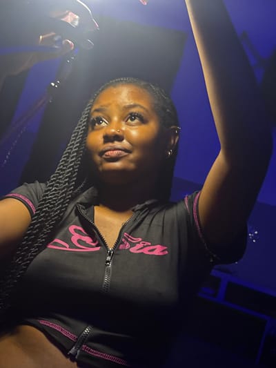 One female subject, appearing to be of African descent, is shown in a medium close-up, gazing upward and slightly to her right. She has dark braided hair, a visible nose piercing on her left nostril, and multiple piercings on her left ear. She is wearing a black, short-sleeved, quarter-zip top with pink lettering, possibly "Ebria," and pink stitching details. A matching lower garment with a zipper is partially visible. Her left hand, extended into the upper left of the frame, holds a small, illuminated device casting light onto her face. Her right arm is raised and extends out of frame to the right. The background is a solid deep blue with faint vertical dark stripes. The scene is illuminated by a strong, localized light source from above left, originating from the device she holds. The location is Gbongan, Nigeria.