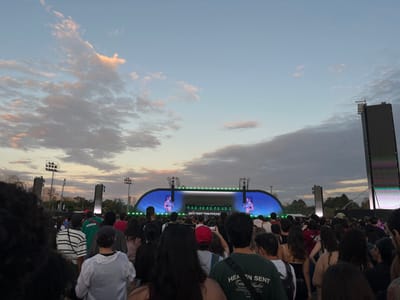 An outdoor concert or festival scene is depicted at dusk in Martínez, Argentina. The image is taken from within a large crowd, looking towards a stage.
The sky overhead exhibits a gradient of light blue at the zenith, transitioning to warmer tones of orange and pink near the horizon, with scattered stratocumulus clouds displaying varied light and shadow.
In the foreground and midground, numerous individuals are visible from behind, comprising a dense audience primarily facing away from the viewer towards the stage. Hair colors vary, and clothing includes casual attire such as t-shirts (one green with "HEAVEN SENT" visible), hooded sweatshirts, and a striped long-sleeve shirt. Several people are holding up smartphones.
The central feature in the midground is a large stage structure, characterized by a curved, horizontal LED screen displaying a performer. Flanking the main stage are vertical light or speaker towers. To the far right, a tall, rectangular LED screen tower stands vertically. In the background, multiple tall stadium-style light poles with floodlights are visible, along with distant trees and some faint urban structures. The overall environment suggests a large open-air venue.
