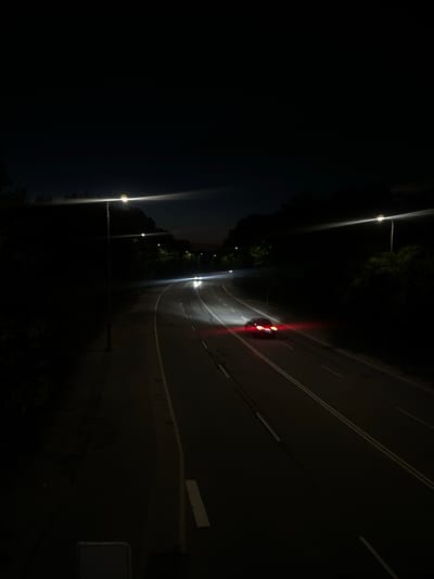 The image captures a dark, nighttime scene of a road, likely a highway or a main thoroughfare, with streetlights illuminating the asphalt. In the middle distance, a car with its red taillights illuminated is driving away from the viewer, creating streaks of light due to motion blur. Other vehicles with headlights are also visible further up the road. The road is bordered by dark trees and foliage on both sides, suggesting a semi-urban or suburban environment. The sky above is a deep, dark blue, almost black, with a faint glow near the horizon indicating the presence of distant lights or the lingering twilight. The overall impression is one of a quiet, possibly deserted stretch of road at night.