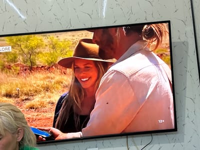 The image displays a television screen showing a scene from what appears to be a documentary or reality show set in an arid landscape with sparse vegetation. On the left side of the screen, the word "LORE" is visible in a white rectangle. In the foreground of the television image, a woman wearing a wide-brimmed hat is smiling. She is positioned next to a man whose face is partially obscured, but he is wearing a light-colored shirt. A rating of "12+" is displayed in the bottom right corner of the television screen.
In the foreground, outside the television screen, a portion of a person's head and shoulder with blonde hair is visible, wearing a green shirt. This person is holding a blue smartphone, with the screen facing towards the television. The wall behind the television has a marbled or textured pattern. The overall scene appears to be captured from a viewing angle within a room, with a cable extending from the bottom right of the television. The location is specified as the city of Benkovski, Bulgaria.