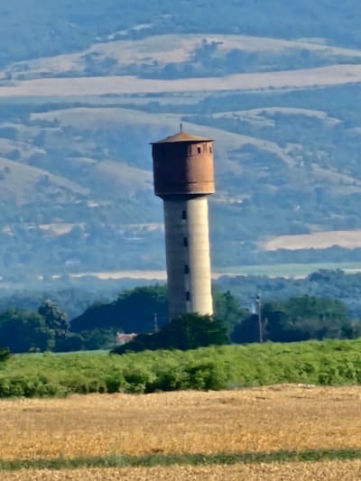 This image shows a tall, cylindrical water tower with a reddish-brown, domed top and circular windows along its white shaft. The water tower is situated in a rural landscape, likely near the village of Zagortsi, Bulgaria, given the context. In the foreground, a field of dry, yellowish vegetation, possibly harvested crops, is visible. Behind the water tower, there is a lush green strip of foliage, and further in the background, rolling hills with scattered vegetation and distant hazy mountains can be seen. The sky is a pale blue. The scene depicts a solitary man-made structure amidst a natural, agrarian environment.