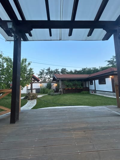 The image captures a view of a backyard in Voyvodino, Bulgaria, from a wooden deck. The foreground is dominated by the wooden planks of the deck, with a dark wooden support post on the left. Beyond the deck, a well-maintained lawn stretches out towards a house with a red-tiled roof. To the left of the lawn, a stone pathway leads to a rustic well with a red roof. The house, partially visible on the right, has white walls with stone detailing at the base and wooden accents. Several outdoor lights are visible, some illuminating the pathway and the house exterior. Trees and foliage surround the property, and the sky is clear and light blue, suggesting it is either early morning or late evening. There are no people or animals visible in the image.