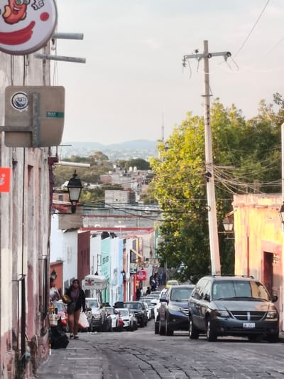 The image depicts a narrow, cobblestone street in Querétaro, Mexico, with buildings of various colors and heights lining both sides. The street slopes downward and is filled with parked cars, including a grey minivan prominently positioned in the foreground. A utility pole with numerous electrical wires stands to the right of the street, adjacent to a building with a yellow-painted facade. A woman carrying bags walks along the left side of the street. In the background, there are rooftops and distant hills visible under a hazy sky. Signs and other urban fixtures are attached to the buildings, contributing to the overall atmosphere.