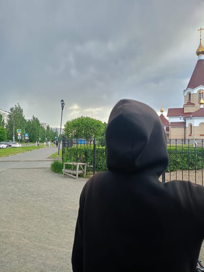 The media file shows a person wearing a black hoodie, seen from behind, standing on a gravel path. In the background, a street runs parallel to a sidewalk where several people are walking. Trees line the street and sidewalk, and multi-story apartment buildings are visible further in the distance. To the right of the person and the path, there is a black metal fence, beyond which are manicured bushes and a wooden bench. Further to the right, a portion of a light-colored brick building with a red roof and a golden dome is visible, suggesting it is a church or religious building. The sky is overcast and appears to be a cloudy day, possibly late afternoon given the subdued lighting. The general atmosphere is calm and ordinary, with no specific event immediately apparent beyond people going about their daily activities in an urban environment. The setting appears to be a residential area in Petrozavodsk, Russia, indicated by the architectural style and the presence of typical urban infrastructure. There are road signs visible, including a pedestrian crossing sign and a speed limit sign showing "20".
