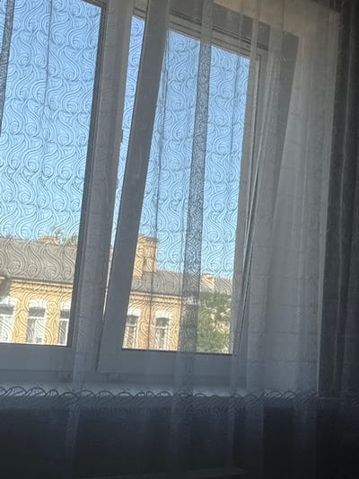 The image captures a view through a window covered by translucent striped blinds, revealing a building across the way. The blinds are made of a sheer fabric with an intricate, wave-like pattern. They are adjusted to allow light to pass through, creating horizontal bands of light and shadow. Through the open sections of the blinds, the sky appears bright blue, suggesting a clear day. The building opposite is a multi-story structure with a light yellow facade. Its windows are visible, and the sunlight casts shadows on its surface. The overall impression is one of daytime, with pleasant weather, seen from an indoor perspective looking outwards. The scene is calm and ordinary, depicting a typical urban or suburban view. There is no visible text in the image.