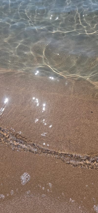 The image shows a close-up view of shallow, clear water washing over a sandy beach. Sunlight refracts through the water, creating shimmering patterns and highlights on the wet sand. The sand is a light brown color and appears fine-grained. In the lower part of the image, a small wave is breaking, creating a line of white foam and bubbles on the sand. The water is calm and transparent, allowing the sandy bottom to be clearly visible. The scene is a natural environment, likely a beach or shoreline. The location is identified as the city of Sary-Oy, Kyrgyzstan. There are no people or animals visible in the image.