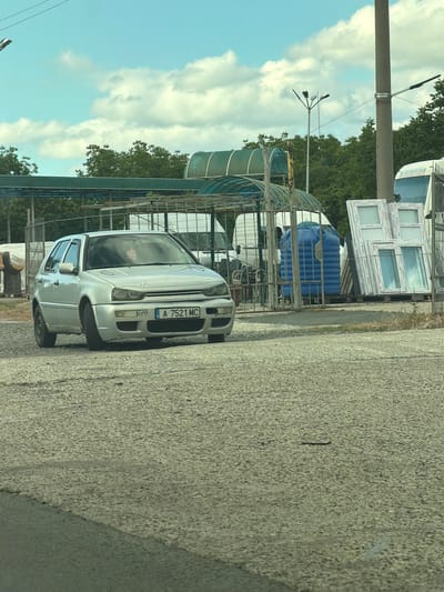 The image shows a silver Volkswagen Golf parked on a paved surface. The car has a license plate that reads "A 7521 MC". In the background, there are stacks of white window frames, a large blue cylindrical tank, and a metal fence structure. Trees are visible behind the structures, and the sky is partly cloudy. The scene appears to be outdoors, possibly in a storage yard or industrial area. The time of day is likely daytime, given the bright sky and shadows. There is no clear indication of the weather beyond the presence of clouds. The overall setting seems to be in Aytos, Bulgaria, as provided by the location context. No people are visible in the image.