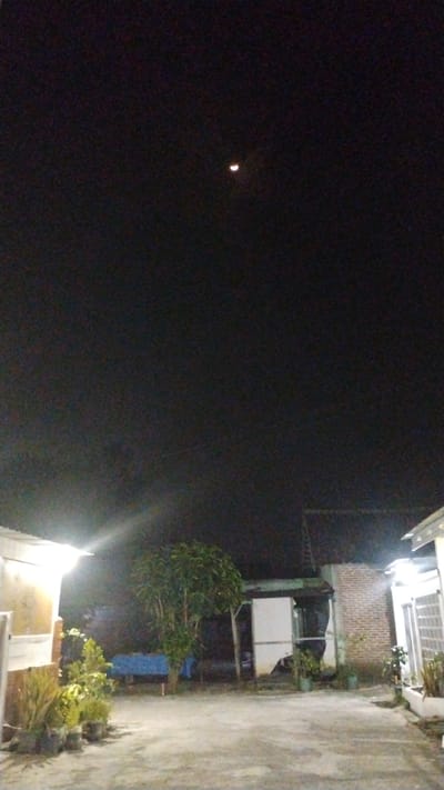 The image depicts a nighttime outdoor scene, likely in a residential area of Banjarsari, Indonesia. A crescent moon is visible in the dark sky. In the foreground, a concrete surface suggests a courtyard or driveway. Several potted plants are arranged near a building on the left, which is illuminated by a bright light. To the right, another building with brick and white facade elements is partially visible, also featuring outdoor lighting. A medium-sized tree stands in the middle ground, with other structures and foliage visible behind it. The overall impression is of a tranquil, slightly dimly lit residential setting at night.
