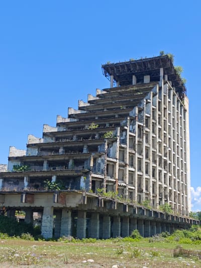 The image displays a large, multi-story, abandoned concrete building under a clear, vibrant blue sky. The structure exhibits a striking architectural design, with its left side characterized by a dramatic stepped or terraced profile where each upper floor progressively recedes. These open ledges and balconies are heavily overgrown with various green plants and bushes, suggesting a prolonged period of disuse. The right side of the building features a more uniform, vertical facade composed of a grid of numerous empty rectangular window or door frames.

The building's concrete surfaces show significant signs of weathering, crumbling, and decay, with some areas appearing to have exposed rebar. The top section reveals an exposed, possibly unfinished roof structure, also with vegetation growing on it. The main body of the building is elevated above the ground by a series of robust concrete pillars or stilts.

The foreground consists of unkempt green grass, weeds, and scattered small rocks and pebbles, with a few small purple wildflowers visible. The scene is brightly lit by what appears to be mid-day sun, indicating clear and sunny weather. There is no visible human activity or text. The image evokes a sense of desolation and natural reclamation of a once ambitious, now decaying, structure located in Warcha, Abkhazia.