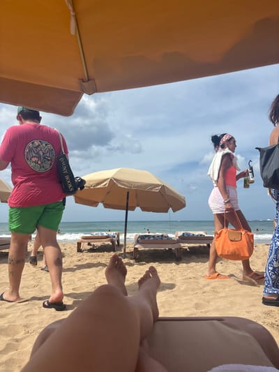 The image captures a beach scene in San Juan, United States, from the perspective of an individual reclining on a beige lounge chair, with their bare legs and feet visible in the foreground. Blue nail polish is discernable on some toes. An orange beach umbrella canopy extends across the top of the frame, providing shade.
In the mid-ground, a sandy beach is populated by individuals and beach furniture. A male subject, seen from behind, walks from left to right. He wears a red t-shirt with a circular graphic depicting a bat and foliage, green shorts, and dark flip-flops. A black tote bag with "DAUNT BOOKS" written vertically in white letters is slung over his left shoulder. Tattoos are visible on his lower legs.
To the right, a female subject also walks from left to right. She wears a coral-colored top, light-colored patterned shorts, and orange slide sandals. A white towel is draped over her shoulders, and a patterned headband adorns her head. She carries two beverage bottles in her left hand and a large orange tote bag in her right hand.
Further back, a tan beach umbrella stands upright on the sand, surrounded by several empty lounge chairs. The ocean extends beyond, displaying gentle waves. Distant, indistinct figures are visible wading or swimming in the water. The sky above is partly cloudy.
