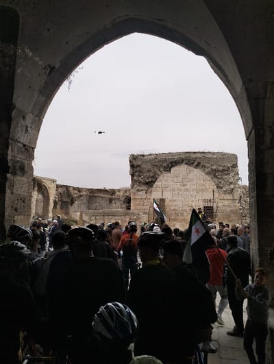 A crowd of people, many wearing bicycle helmets, gathers in what appears to be the entrance of an old, stone structure. Two Syrian flags are visible amidst the crowd. In the sky, a small helicopter is flying in the distance. The stone entrance is arched and leads to a larger open area containing more stone ruins. A young boy is standing towards the right side of the image. The overall scene appears to be in a historic or archaeological site.