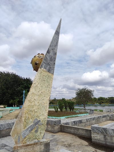 The image displays an outdoor scene featuring a large, ornate monument under an overcast sky. The original image appears to be rotated 90 degrees counter-clockwise.
The central focus is a tall, triangular monument, rising diagonally from the lower right towards the upper left of the frame (when viewed correctly). The monument is constructed from multiple distinct materials:
* The top, pointed section is dark grey or black, with a textured, striated, or veined pattern, resembling polished stone like granite or marble.
* Below this, the mid-section is lighter, appearing light beige or yellowish. This surface is highly textured, filled with numerous imprints that strongly resemble human handprints, suggesting it might be a wall or column featuring a collection of hand casts or impressions.
* Attached to the monument, slightly below the transition from the dark grey to the handprint section, is a golden, spherical object. This sphere also has a rough, textured surface.
The base of the monument and the immediate surrounding structures are made of grey, block-like stone, similar in appearance to the top section of the monument, possibly granite or marble. These structures include what looks like steps or decorative barriers.
In the background, under a sky filled with large, fluffy white and grey clouds, there is a body of water visible, possibly a lake or river, with a faint blue edge or barrier along its shore. Beyond the water, a line of green trees and foliage is visible. A prominent, large tree with a dense canopy stands in the mid-ground, towards the left of the frame. The ground in the foreground appears to be a mix of paved areas and some dirt or dry vegetation.
The lighting is diffuse and soft, consistent with an overcast daytime, and there are no harsh shadows. No people are visible in the frame, and there is no discernible text. The overall impression is that of a public park or memorial site.