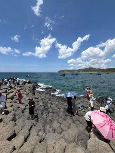 The image depicts a sunny day at a rocky coastal area, possibly in Phú Lương, Vietnam. A group of people are gathered on a formation of dark, hexagonal basalt columns that extend into the sea. The sea is a vibrant blue-green, with waves gently lapping against the rocks. Several small boats are visible on the water in the middle distance, and a hilly coastline can be seen in the background.
The people are dressed in casual summer clothing, and some are holding umbrellas to shield themselves from the sun. A few individuals are posing for photos, while others are walking or standing around, enjoying the view. One person in the foreground is crouching down, and another is holding a pink umbrella with red graffiti on it. The overall atmosphere appears relaxed and tourist-oriented.