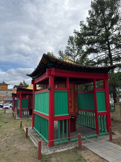 A low-angle shot depicts a series of small, elaborately decorated red and green structures under a cloudy sky in Ulaanbaatar, Mongolia. The structures feature ornate roofs with detailed carvings and green latticework. The red paint is a primary color, contrasting with the light green. Each structure has a painted interior and is linked by a rope fence with evenly spaced wooden posts. The background includes a tall tree and modern buildings, contrasting the traditional structures. Pedestrians are visible in the background, and the surrounding ground is a mix of paving stones, cobblestone, and grass.