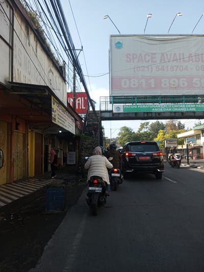 The image shows a street scene in an urban environment. There are buildings on the left side of the frame, with storefronts and an overhead canopy. A pedestrian is walking on the sidewalk near the storefronts. An overpass spans the road with an advertisement visible on the structure. The road has vehicular traffic, including a black SUV and several motorcycles. A person wearing a white jacket is driving one of the motorcycles. Electrical wires are visible overhead. The environment seems to be a mix of commercial and residential areas. The overpass sign has text on it. The street appears to be in Indonesia, based on the license plates visible on the vehicles. The license plates are in the format used in Indonesia. "CIPANAS" is visible on a sign near the overpass.