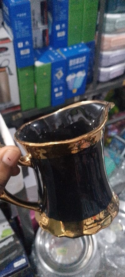 A human hand, located in the lower-left foreground, holds a small, black, glossy ceramic creamer or pitcher by its ornate gold-colored handle. The creamer features a contoured body, a defined spout, and gold trim along its rim and base. A small, dark mark is visible on the tip of the thumb holding the handle. The blurred background depicts an indoor commercial setting, likely a retail space within Okere-Urhobo, Nigeria. Visible background elements include stacked retail packaging: predominantly blue boxes, some with white text and graphics (e.g., a cloud design), interspersed with green boxes and vaguely discernible lighter-colored containers on the right. Artificial illumination is evident, casting reflections on the creamer's surface.