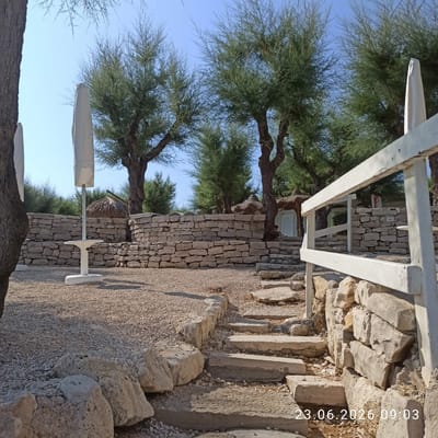 The image depicts an outdoor scene featuring a pathway leading up to a stone wall, with trees and foliage in the background. The pathway is made of uneven stone slabs and is bordered by large rocks and gravel. A white railing runs along the right side of the pathway. Behind the railing, a stone wall is visible, constructed from roughly hewn stones. In the background, several trees with sparse foliage are present, and beyond them, a structure with a thatched roof and a white door is partially visible. Two white, covered umbrellas or flags stand upright to the left of the pathway. The overall environment appears to be a well-lit, outdoor area with a natural, slightly rustic aesthetic. The image contains a timestamp in the lower right corner: "23.06.2026 09:03". There is a person seated on the steps to the right of the thatched roof structure. This location appears to be a beach or coastal area, possibly a resort or park, given the presence of umbrellas and the natural landscaping.
