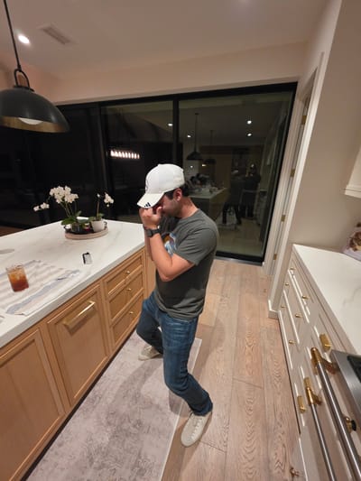 The image depicts a man standing in a modern kitchen or open-plan living space, viewed from a slightly elevated perspective. The man is positioned towards the right side of the frame, leaning slightly forward and looking down and to his left. He is wearing a dark olive green short-sleeved t-shirt, blue jeans, and white sneakers. A white baseball cap with an "Adidas" logo is worn backward on his head, and a dark-colored smartwatch is visible on his left wrist.

The setting features a bright, clean aesthetic with light-colored wooden flooring and a patterned light-toned rug. To the man's right, a long kitchen island or set of base cabinets made of light wood with gold bar handles and smaller gold knobs is visible. The countertop is a white marble-patterned surface. On the countertop, there is a potted white orchid plant, a clear glass containing an amber-colored drink, and a striped towel. A small white object with "PAX ME" visible text is also present on the counter.

Behind the man, a large, dark-framed glass door or window stretches across the wall, showing reflections of indoor lights and indistinct figures. This suggests it is evening or nighttime outside. To the far right, a set of white upper cabinets with gold handles and a marble-patterned countertop incorporates a stainless steel oven or stove. A dark, modern pendant light fixture hangs from the ceiling in the lower left portion of the image. The overall scene conveys a contemporary residential interior, possibly in Coronado, United States of America.