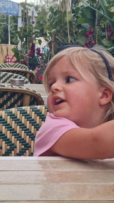 A photograph captures a young, blonde-haired girl with a black headband, wearing a pink shirt. Her expression is lively, with an open mouth and eyes looking slightly to the left. She leans against what appears to be a table or counter with a light-colored, vertically-striped surface.

Behind her are woven chairs with a green and beige pattern. In the background, foliage and flowers are visible, along with a sign in Hebrew, indicating the presence of parking nearby. The location is Kiryat Gat, Israel, as indicated on the sign.