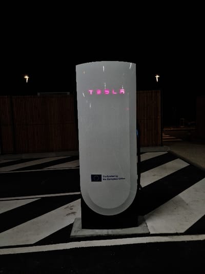 The image displays a Tesla Supercharger V3 station positioned centrally in an outdoor parking environment at night in Les Tourrettes, France. The tall, light gray charging unit features a magenta-illuminated "TESLA" logo near its curved top. Below the logo, a graphic shows the European Union flag next to the text "Co-funded by the European Union." The unit is mounted on a dark, rectangular base, which rests on a lighter concrete slab. The ground in the foreground is marked with prominent black and white diagonal stripes on an asphalt or concrete surface. Behind the charging station, a dark wooden slatted fence or wall extends across the midground. The background is a dark, clear night sky, with two distinct, distant yellow-white light sources visible in the upper left and right portions of the frame. No individuals, vehicles, or specific actions are present in the scene.