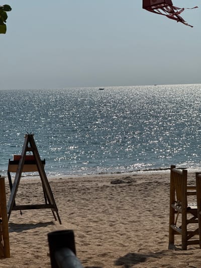 A daytime scene depicts a sandy beach adjacent to a body of water in Phra Ae, Thailand. The sky is clear with a slight haze at the horizon. The water surface, likely a sea or ocean, exhibits extensive glinting from direct sunlight, creating numerous bright, reflective points. The sandy beach in the foreground and midground shows fine texture with ripples and slight indentations.
In the left midground, a dark, weathered wooden A-frame structure stands on the sand, with a horizontal beam suggesting a support or display. Partially visible in the right midground are components of two light-colored wooden chairs or stools, possibly made of bamboo, positioned facing the water. A dark, out-of-focus object, potentially part of another structure, is visible in the bottom foreground. Shadows are cast onto the sand from unseen objects to the right. From the top right, a reddish-brown, box-shaped kite with multiple streamers hangs in the upper sky. A few dark green leaves are partially visible from the top left corner, suggesting nearby foliage.
On the water in the distant background, two small, dark boats are visible. One is positioned slightly left of center, and the other is further right near the horizon line. No individuals are present or engaged in any direct actions. The scene is static, under bright, direct sunlight.