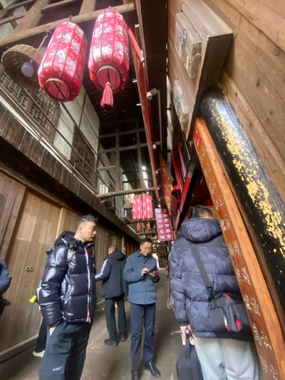 The photo captures an indoor scene, likely a street in a historical district. Red lanterns hang from the ceiling above wooden structures. Several people are seen walking through the area, with some wearing puffy jackets. There are signs with Chinese characters displayed. The space has an old, traditional atmosphere.