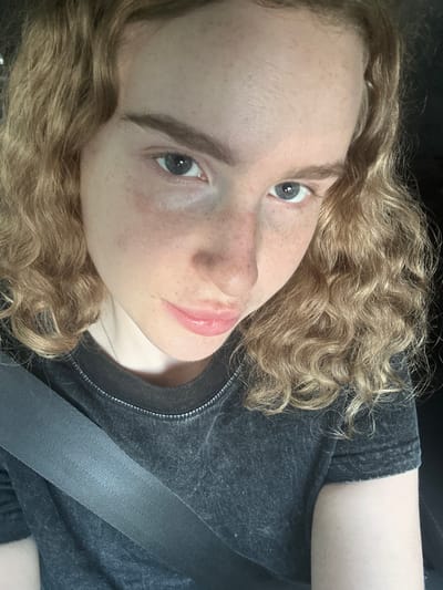 A young person with curly blonde hair, fair skin, and freckles is taking a selfie in a car, as indicated by the presence of a seatbelt. The person is wearing a dark grey t-shirt. The background is dark and out of focus, suggesting the interior of a vehicle. The image does not contain any information to suggest a location in Vasilievo, Russia.