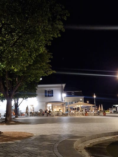 The image depicts an outdoor seating area of a restaurant or cafe in Überlingen, Germany, at night. A large tree with dense foliage is prominently positioned on the left side of the frame, partially obscuring the view of the building behind it. The building appears to be a two-story structure with a white facade and a blue-shuttered window on the upper level.
In front of the building, numerous tables and chairs are set up for outdoor dining, some covered by large, light-colored umbrellas. Several people are seated at the tables, appearing to be patrons of the establishment. The area is illuminated by artificial lights, creating a warm glow.
The ground in the foreground is paved with cobblestones, and a curved concrete structure, possibly a planter or seating area, is visible. The background is dark, indicating nighttime, with some distant lights and a faint horizontal light streak across the sky, likely a lens flare from a light source. A street lamp is visible on the right side of the image. The overall impression is of a lively evening scene in a public space.