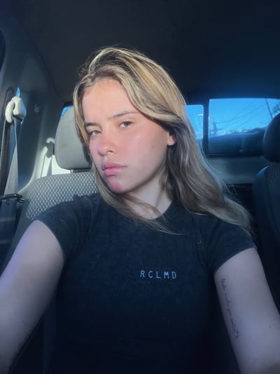 A young female, likely in her late teens or early twenties, is depicted in a close-up selfie taken from within a vehicle. She has long, wavy, blonde and light brown hair, and appears to be looking directly at the camera. A distinct reddish-pink mark is visible on her lower left cheek/jawline. She wears a dark grey, ribbed, short-sleeved top with "RCLMD" printed in white on the upper chest. A vertical script tattoo is visible on her left forearm.
She is seated in what appears to be the front passenger seat of a car or truck, evidenced by the grey fabric seat, headrest, and an unbuckled grey seatbelt fixture to her left. Strong sunlight illuminates her face from the viewer's left. Through the vehicle's rear window, a clear blue sky is visible, along with some green foliage and utility lines, suggesting an outdoor urban or suburban environment during daytime. The image provides no specific visual cues to confirm the location as Santa Rosa, Argentina.