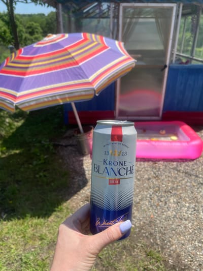 A person's hand with light purple fingernail polish is holding a can of Krone Blanche Biere in an outdoor setting. The can has a white and blue label with gold lettering. Behind the can, a colorful striped umbrella is partially visible, casting a shadow on the ground. In the background, there is a small inflatable pink pool with some yellow objects inside. Further back, a blue structure with a glass door is visible. The environment appears to be a sunny day with green grass and trees. The location is identified as the city of Yasnoe, Russia.