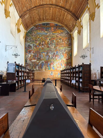 The image depicts the interior of a large, high-ceilinged room, possibly a library, archive, or historic hall, characterized by its impressive architecture and a monumental mural. The room is oriented such that a vast, colorful mural covers a significant portion of the left wall, while rows of dark wooden bookshelves line the right side, receding into the distance.
The mural is highly detailed and vibrant, portraying a dynamic and complex scene. It features numerous figures, including men on horseback, warriors, and other individuals, engaged in what appears to be a historical battle or a significant narrative event. The artwork is rich in color, utilizing shades of red, blue, green, and various earthy tones.
Above the mural, a striking architectural feature is visible: a large, curved wooden structure, possibly part of a vaulted ceiling or a large archway, constructed from horizontal planks with a warm, reddish-brown tone and visible wood grain. Higher up, the walls are painted a light color (white or cream) and are adorned with decorative, yellowish-gold trim or molding. Several rectangular openings, which appear to be skylights or recessed windows, are set into the upper sections, allowing natural light to illuminate the space. A classic wrought-iron chandelier with multiple arms hangs from the ceiling.
On the right side of the room, multiple rows of dark wooden bookshelves or display cases extend far into the background. These shelves are filled with books or other items. Small white signs are attached to the front of many shelves. While most text on these signs is illegible due to distance, one sign clearly reads "NO FOTOGRAFIAS," indicating a no-photography policy. Another sign visible on a shelf includes the word "ESPAÑOL," possibly denoting a language section. Interspersed among the dark bookshelves, or running parallel to them, are long, lighter-toned wooden tables or benches.
In the immediate foreground on the right, a large, dark, polygonal beam or structural element runs horizontally across the lower part of the frame. This beam has visible metallic fasteners (screws or bolts) and appears to be made of dark metal or painted wood.
The floor of the room is covered with reddish-brown tiles, likely terracotta, arranged in a grid pattern. The overall lighting is bright, suggesting daytime, with ample natural light complementing the interior's historical ambiance. No people are visible in the scene. The architecture and the mural suggest a setting with historical or cultural significance, possibly in a Latin American or Spanish colonial context.