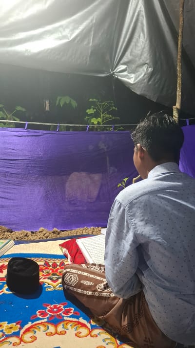 A young man is sitting on a blue and red patterned carpet in what appears to be an outdoor, makeshift shelter. The shelter is covered with a large gray tarp and a purple cloth hangs in the background. In front of the man, a book is open, suggesting he is reading or studying. To his left, a black cap, possibly a peci, rests on the carpet. The ground around the carpet is covered with dirt and sand, with some small plants visible behind the purple cloth. The man is wearing a light blue, pinstriped shirt and a brown sarong. The scene appears to be set at night or in a dimly lit environment, illuminated by an overhead light source. The setting is described as being in the city of Grong-grong, Indonesia.