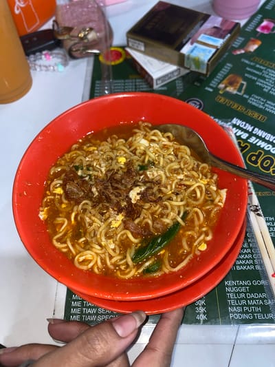 A red bowl of instant noodles, topped with fried onions and a halved egg, sits on a green menu in Mekar Bakti, Indonesia. A spoon rests in the bowl. To the left, an orange bottle and a small pink pouch with gold accents are visible. To the right of the bowl, a pair of wooden chopsticks lies on the menu, which displays various food items and prices in Indonesian currency.