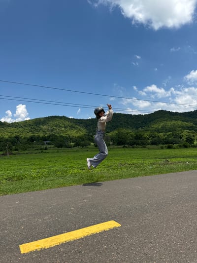A person is captured mid-air, jumping on a paved road with a yellow line. They are wearing a light-colored shirt, blue jeans, and a helmet, with their hands raised in a celebratory gesture. The background features a lush green field, a dense forest covering rolling hills, and a bright blue sky with scattered white clouds. Power lines are visible stretching across the upper portion of the image. The scene appears to be in a rural or natural setting, possibly within the city of Ham Thuan Bac, Vietnam, given the landscape.