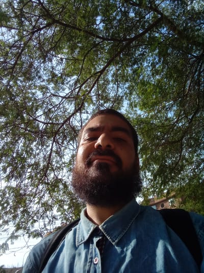A low-angle selfie shows a man with dark hair, a full beard, and a blue denim shirt. He appears to be standing under a tree with dense foliage. In the background, glimpses of the sky are visible through the leaves. Buildings are also visible in the distance. The location is Parroquia Guatire, Venezuela. The man has a backpack on.