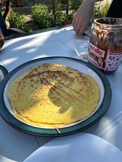The image shows a plate of crepes on a white table, next to a jar of Nutella. A person is using a knife to spread the Nutella. In the background, there are plants and what appears to be a garden. The specific location in Lehchevo, Bulgaria cannot be confirmed from the image.