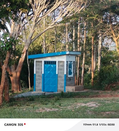 A small, single-room building with a blue door and blue trim stands amid trees and grass. The building is constructed of cinder blocks, painted gray and white, and has a flat, blue roof. A small window is visible on one side. It is surrounded by greenery and various trees, creating a natural setting. Camera information is at the bottom of the frame.