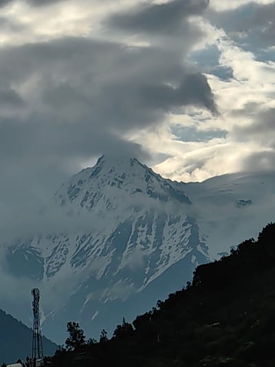 A snow-capped mountain dominates the background, shrouded in clouds. In the foreground, a dark, tree-covered slope descends to the left, where a tall, lattice tower stands. The sky above is filled with dramatic, grey clouds, with patches of sunlight breaking through. The scene appears to be set in the city of Kardang, India.