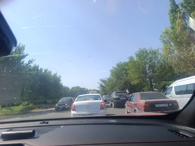 The image depicts a traffic jam on a sunny day in Ötegen Batyr, Kazakhstan. The perspective is from inside a vehicle, looking forward through the windshield. A line of cars is stopped on a two-lane road bordered by trees on both sides. The sky is clear and blue. Visible vehicles include a white Chevrolet sedan in the foreground, a dark red sedan with license plate "KZ 957 JOZ 05" to its right, a dark SUV with license plate "218 AJY 05" further ahead, and a white van behind it. Several other vehicles are visible in the distance. The road appears to be asphalt. The overall scene is one of typical urban congestion during daylight hours.