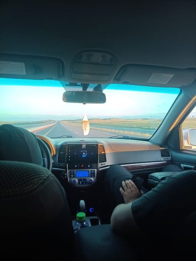 The media file shows the interior of a car driving on a highway in Baynazar, Kazakhstan. The perspective is from the back seat, looking forward. A person's legs are visible in the foreground, with a hand resting on a smartphone. The car's dashboard is prominent, displaying climate control settings and a media player screen with text, although it is not fully legible. A dangling air freshener hangs from the rearview mirror.

Outside the car, a long, straight highway stretches into the distance, bordered by a guardrail and flat, open terrain with some vegetation. The sky is a clear, pale blue, suggesting it is either early morning or late afternoon, likely during daylight hours. The lighting indicates a calm and possibly serene atmosphere. The overall scene captures a moment of travel on a road trip. No specific events or emotions are conveyed beyond the act of driving.