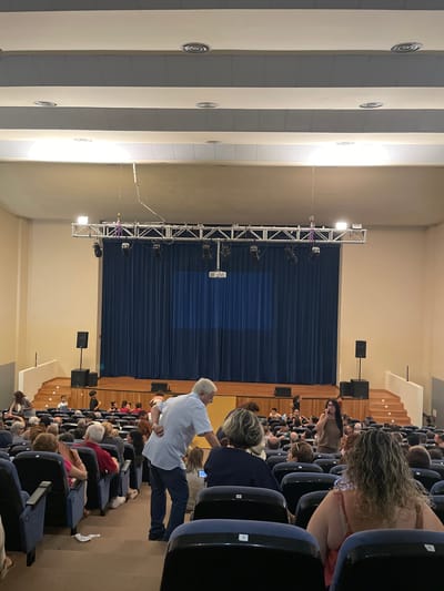 This media file captures an indoor scene within a venue, likely a theater or auditorium. The primary subjects are a large group of people seated in rows of dark blue chairs, facing a stage area. The stage is draped with a dark blue curtain, and above it, a metal truss structure is visible, equipped with various lights and speakers, suggesting a performance or event is about to begin or has just concluded.
A man in a light blue shirt and dark pants is standing and facing away from the audience, towards the stage. He appears to be in motion. Other individuals are visible in the audience, some looking towards the stage, others engaged in conversation or looking at their phones. The lighting suggests it is likely daytime, though the indoor setting makes it difficult to determine precisely. The architecture of the venue is partially visible, with light-colored pillars or walls on the left side of the frame.
The atmosphere appears to be one of anticipation or gathering for an event. The seating arrangement and stage setup are typical of a performance space. No specific text is visible in the image. The location context of Sagunto/Sagunt, Spain, implies this venue is situated in that region.